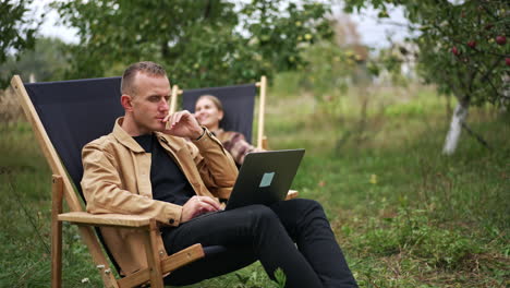 thoughtful male freelancer looking at the screen of his laptop. lady sitting behind having phone conversation. remote work concept.