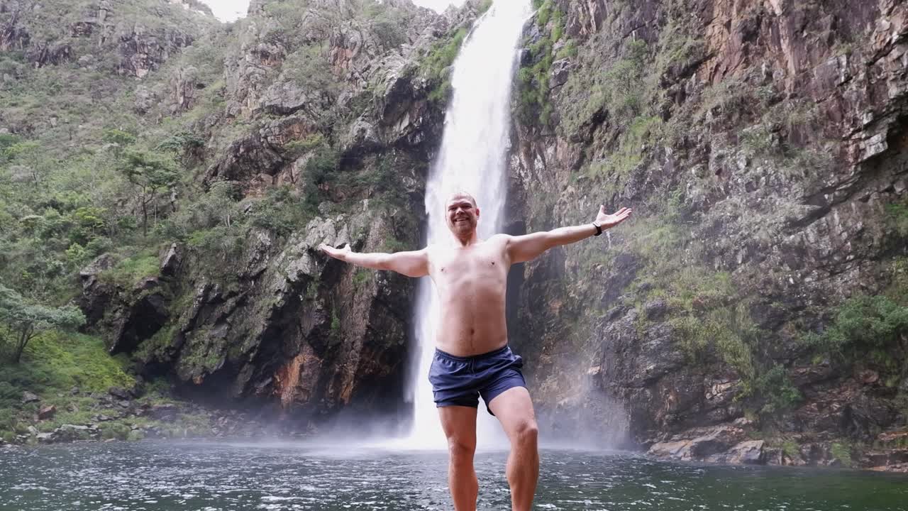 Young blond man poses below Fundao Waterfall in remote Brazil landscape