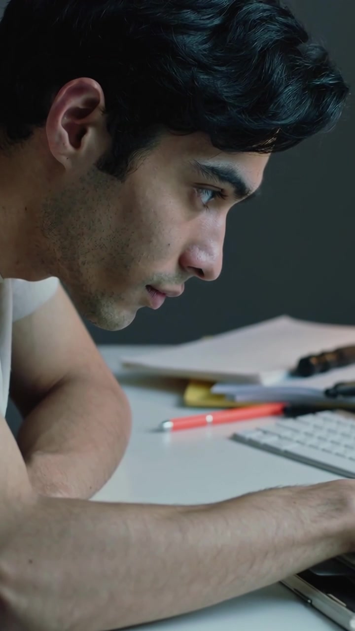 Video verticale: Uomo appoggiato in camicia bianca che raggiunge la tastiera, digitando per finire gli appunti su una scrivania poco illuminata.