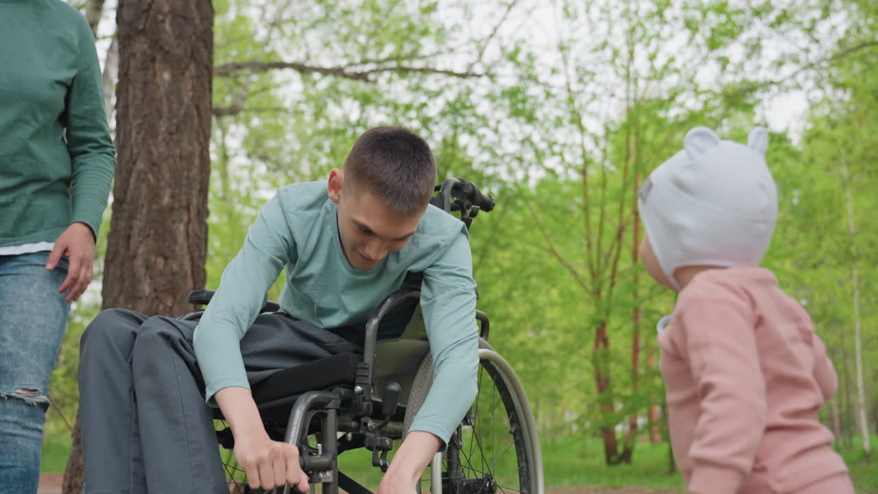 Park White Man Wheelchair Reaches Toddler, Caregiver Nearby Guiding Child Steps, Gentle Touch Exchange, Spring Greenery, Inclusive Bonding Moment, Playful Outreach And Family Support