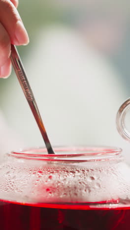 una mujer agita el té en una tetera de vidrio con una cuchara de primer plano. una mujer prepara una bebida herbal caliente para el desayuno en la mesa. proceso de elaboración de una bebida aromática de hibisco