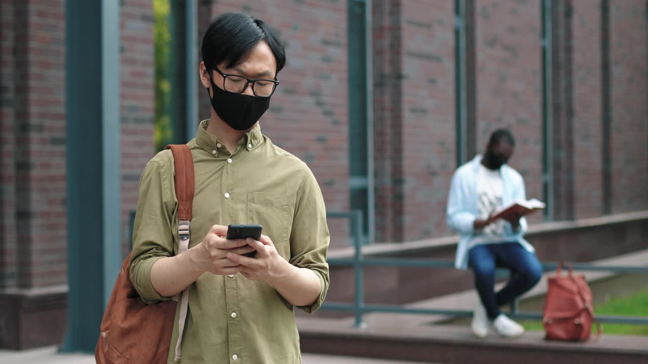 Asian student wearing glasses and facial mask using his smartphone near the college