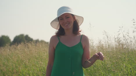 lady in green dress and white sun hat stands smiling while gently touching tall barley in open countryside under bright sunlight with peaceful expression and natural background