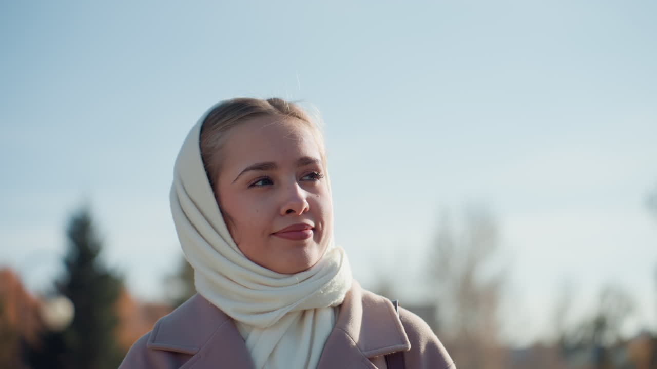 Caucasian woman wearing white scarf standing in sunlit autumn park, contemplative expression, soft coat, gentle breeze, shallow depth, warm tones, closeup portrait, reflective mood, morning light