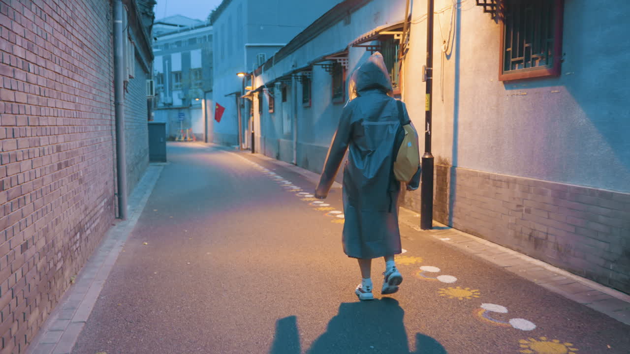 物思いにふける夜の都会の散歩。フード付きコートをまとった穏やかな女性が、静かな都会の路地を進む。狭い通りを、黄色のバックパックを背負った女性が、柔らかい街灯の光の下を静かに足を進めている。