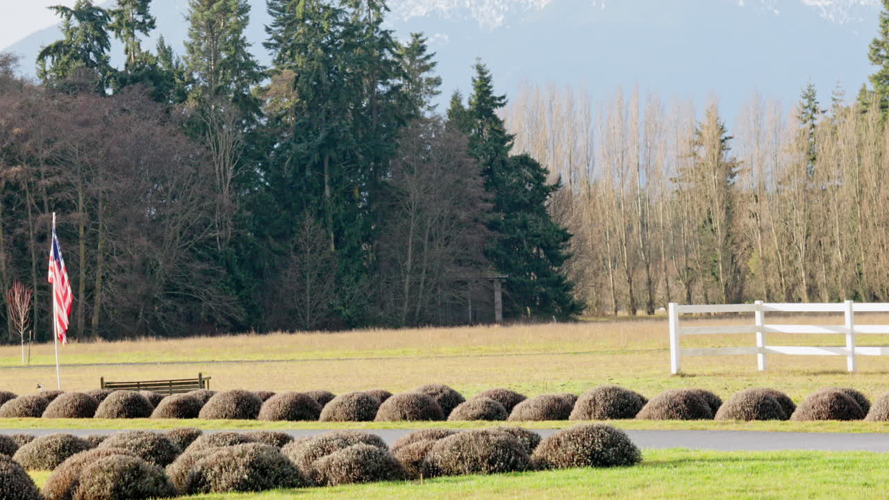 sartén a la izquierda a través de la cerca blanca con la bandera de ee.uu. al lado de la carretera en un poste con árboles en el fondo
