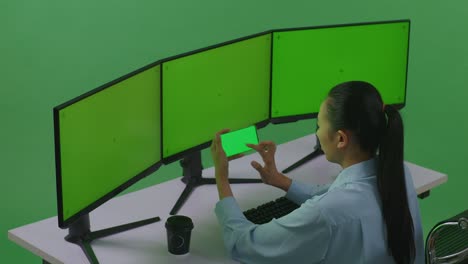 vista lateral de una mujer asiática usando un teléfono móvil y varios monitores de computadora con una maqueta de pantalla verde en un estudio de pantalla verde