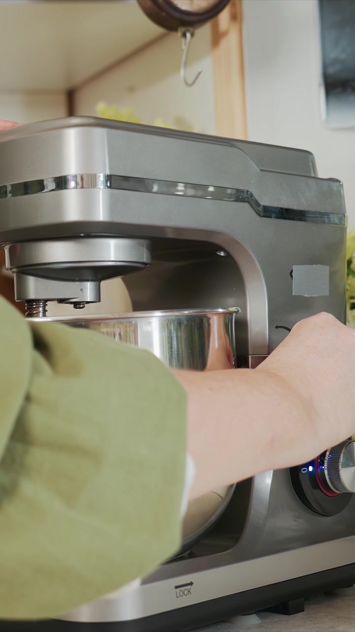 Woman wearing green sleeve adjusts and operates electric. Scene shows close up interactions between hands mixing. White ceramic container is visible on countertop as floral decor