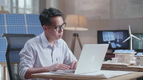 side view of unsatisfied asian man working with laptop computer with model of a small house with solar panel