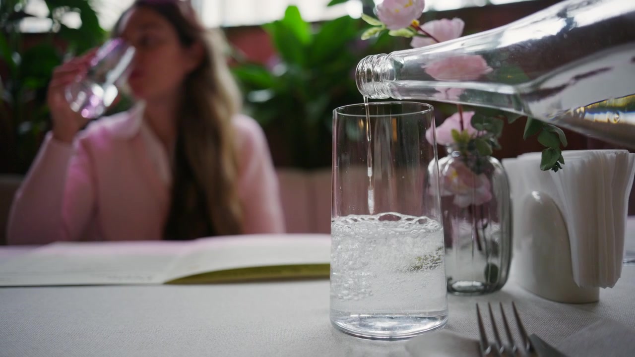 Nahaufnahme von Wasser, das aus einer Glasflasche in ein klares Trinkglas auf einem Restauranttisch gegossen wird. Im unscharfen Hintergrund trinkt eine Frau Wasser.