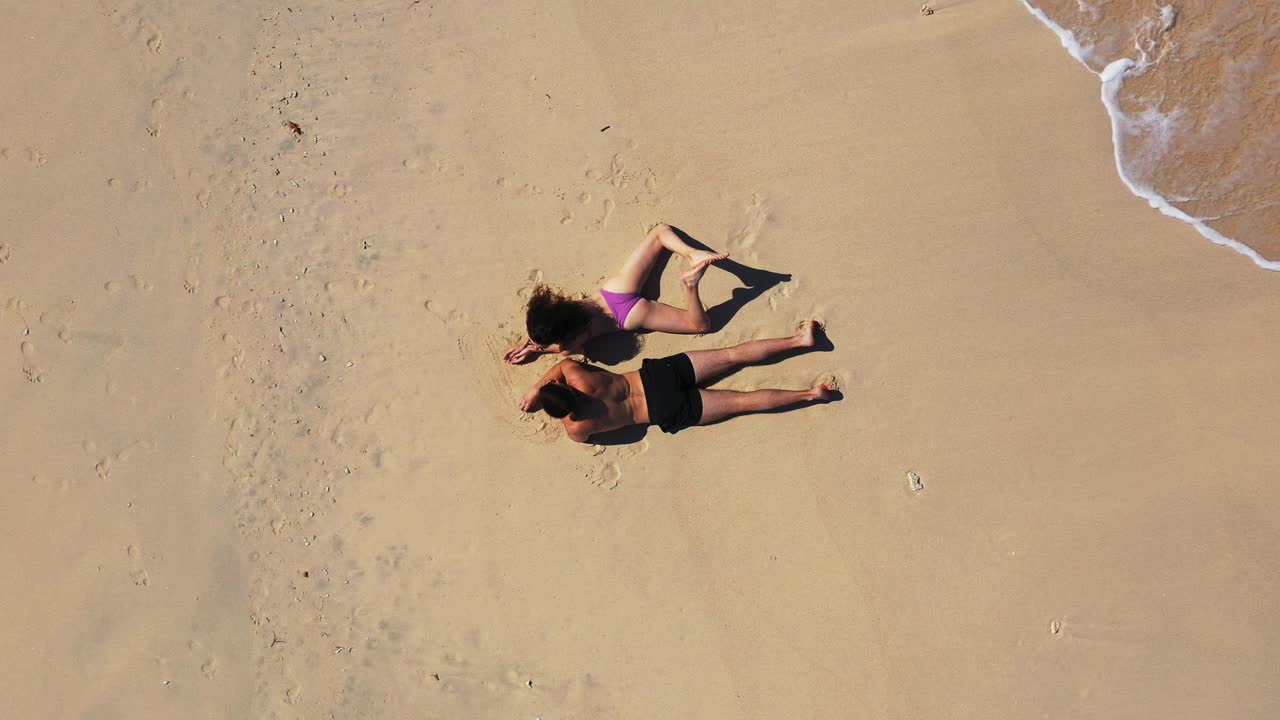 Couple in swimsuit lying on sandy exotic beach writing in wet sand their names washed by ocean flows, copy space