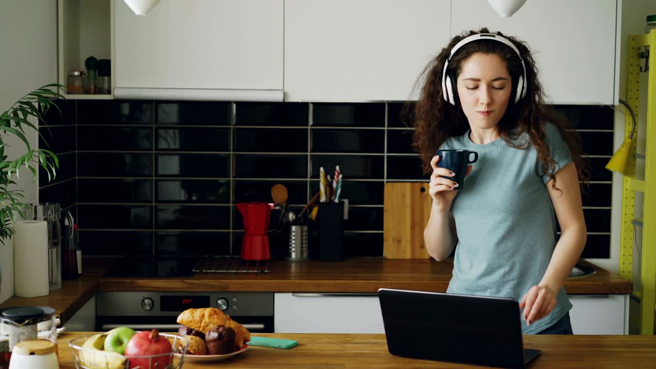 joven hermosa mujer caucásica rizada con auriculares bailando en la cocina cerca de la mesa frente a la computadora portátil, imprimiendo algo, tomando el teléfono y enviando mensajes de texto