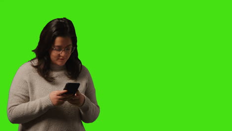 fotografía de estudio de una mujer joven usando un teléfono móvil para enviar mensajes de texto y desplazarse por internet de pie contra un fondo de pantalla verde 1