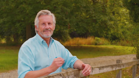 Portrait-Of-Smiling-Casually-Dressed-Mature-Or-Senior-Man-Leaning-On-Fence-On-Walk-In-Countryside