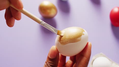 manos pintando huevos blancos usando pincel con pintura dorada sobre fondo púrpura