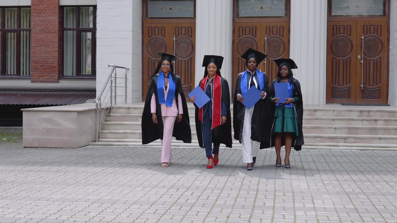 Happy graduates of a university or college of African American nationality with blue diplomas in their hands walk forward to the camera from the entrance to the university