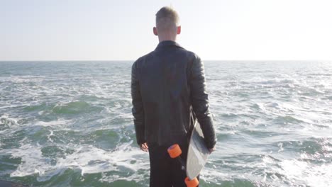 Young-man-in-black-leather-jacket-holds-his-longboard-by-the-sea