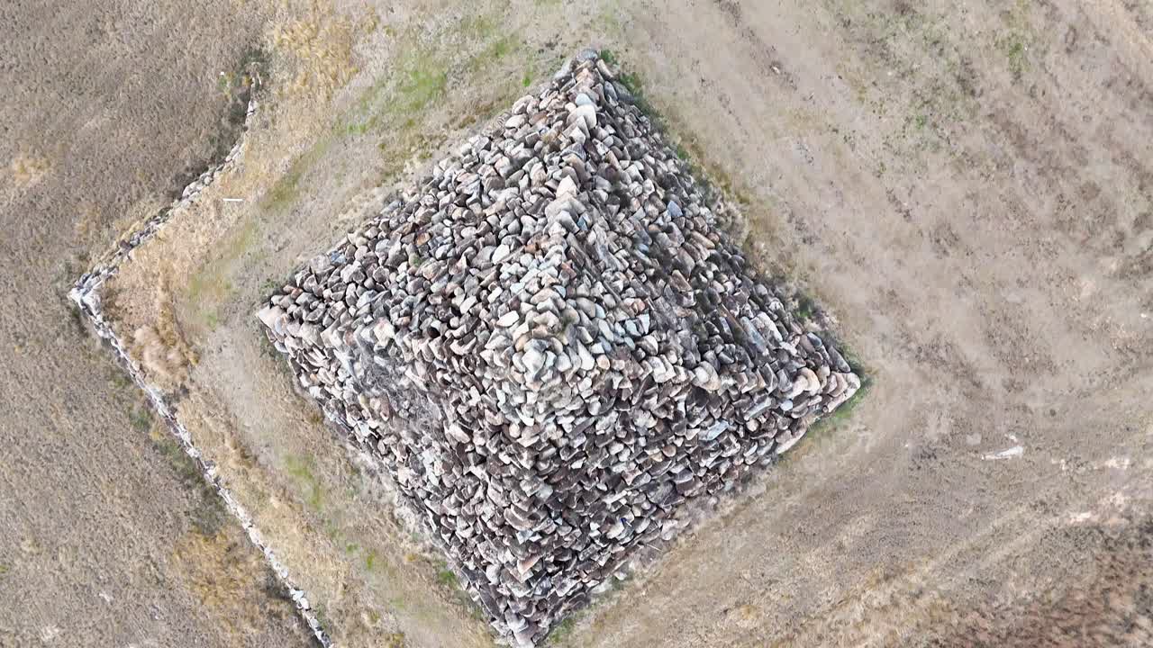 A drone camera smoothly ascends above a large stone pyramid structure in a dry, open field near Ballandean, Queensland, under diffuse daylight