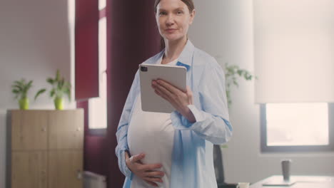 pregnant woman posing at camera while touching her belly and holding a tablet in the office