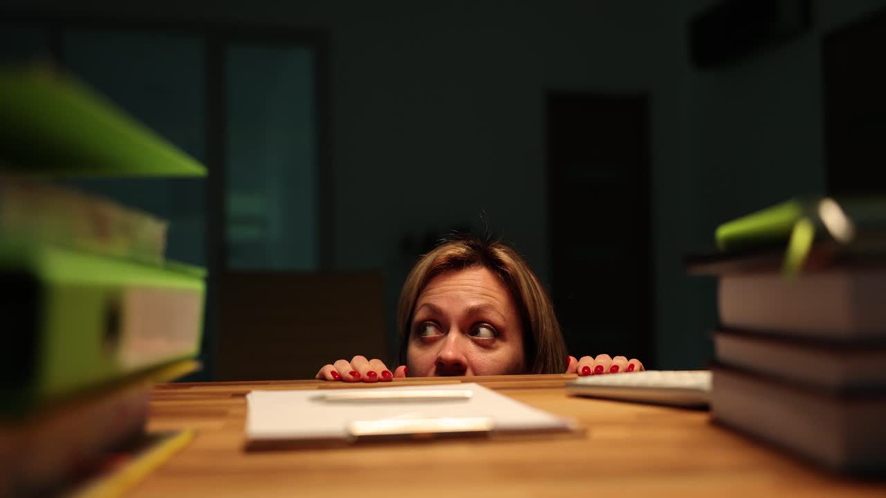 Woman peeking over a desk, looking scared or startled in a dimly lit room