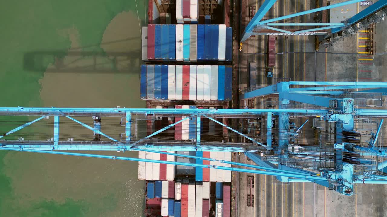 gantry cranes moving containers from a docked cargo ship, illustrating global shipping and supply chain logistics. Smooth aerial view vertical bird's eye view drone