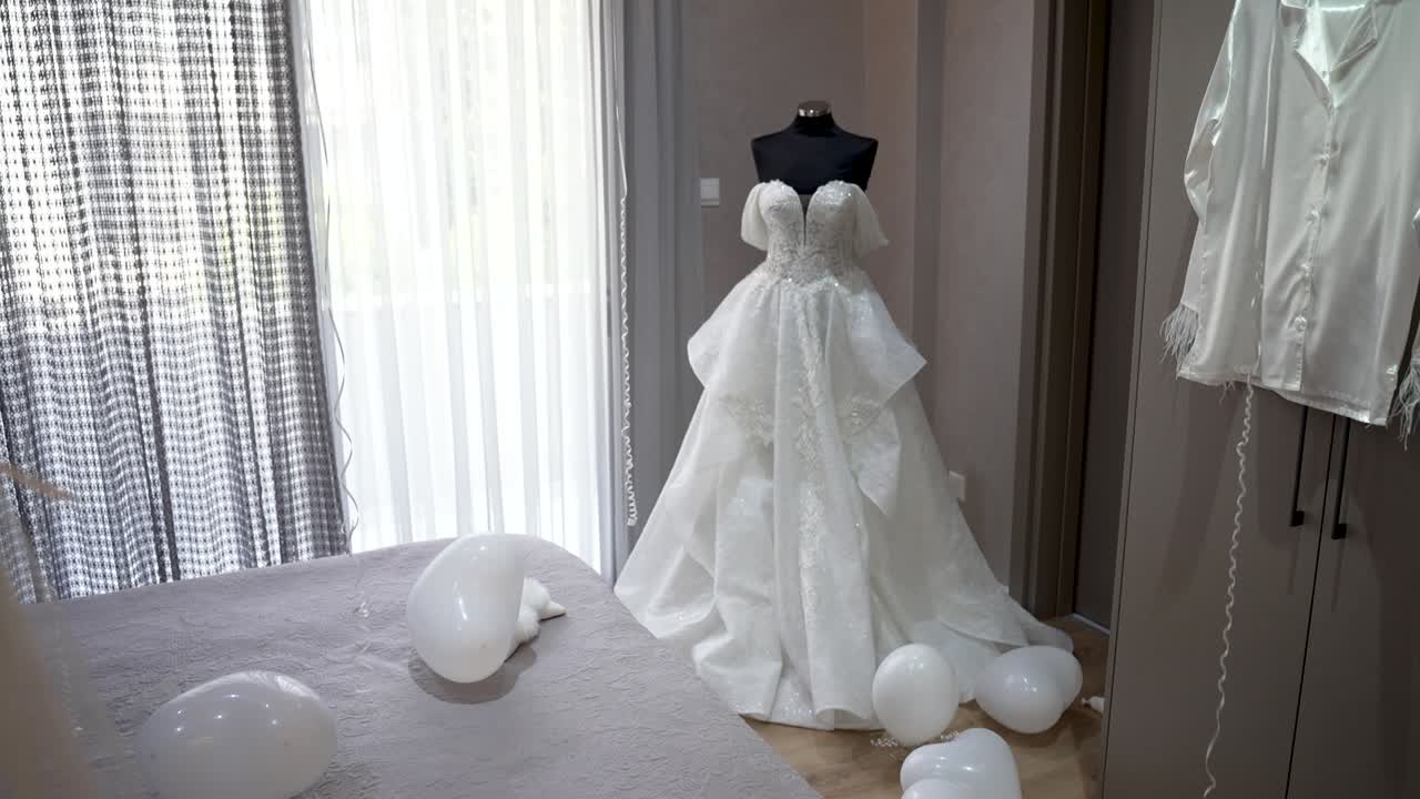 A bride gets ready for her wedding day in a bright room. A white wedding dress is on display, surrounded by simple decorations like balloons. The light shines through the windows