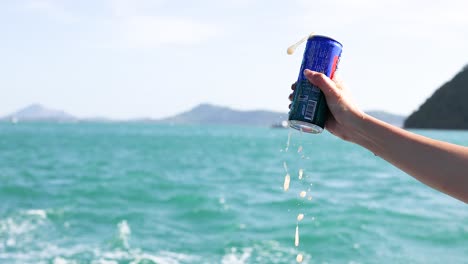 una mano agita una lata de refresco, haciendo que hume y se derrame contra un paisajístico fondo oceánico