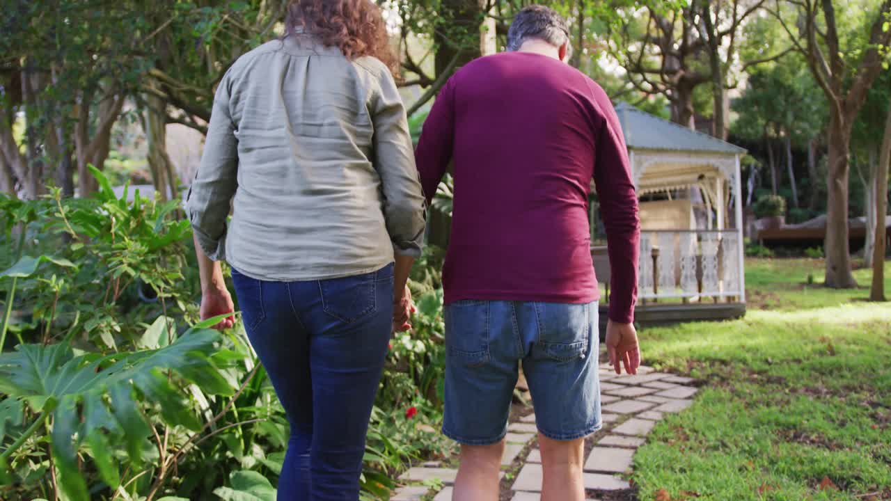 vista trasera de una feliz pareja caucásica mayor cogida de las manos, caminando en el jardín