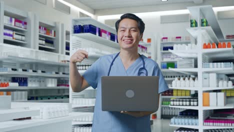 médico masculino asiático con estetoscopio sonriendo y apuntando a una computadora portátil en la mano mientras está de pie en la farmacia