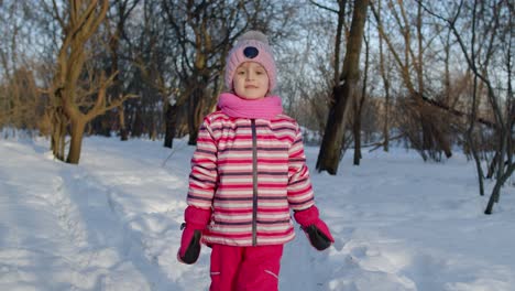 Child-girl-walking-on-snowy-road,-fooling-around,-smiling,-looking-at-camera-in-winter-park-forest