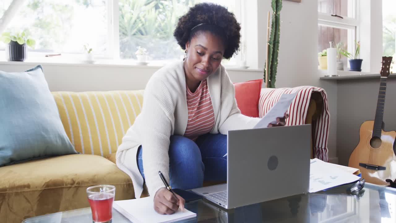 Heureuse femme afro-américaine travaillant à la maison, gérant des documents et utilisant un ordinateur portable, au ralenti