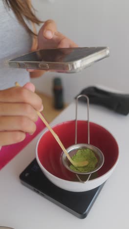 person sifting matcha powder into a bowl while holding a phone for tea preparation
