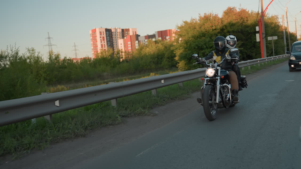Motorcycle rider with passenger drives along roadside guardrail wearing helmets and leather jackets, car coming from behind, lush green trees in background