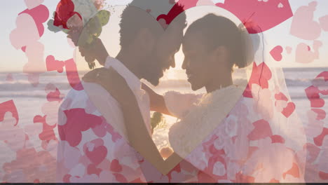 animation of hearts over happy diverse just married couple embracing on beach by sea