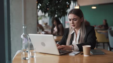 beautiful, sad young woman sits in a cafe in business attire, saddened by what she sees on her laptop, grasping her head