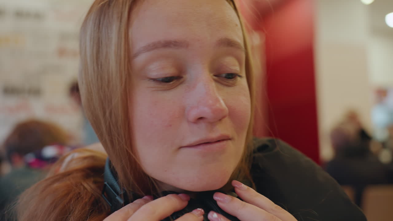 White woman in busy cafe student adjusts headphones while scanning crowd, tense expression, red interior backdrop, candid closeup portrait conveying vigilance and concern about snatcher risk