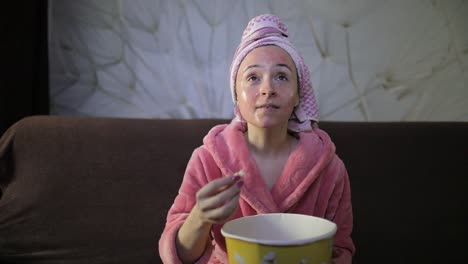 woman watching a late night movie at tv, eating popcorn. bathrobe, facial mask