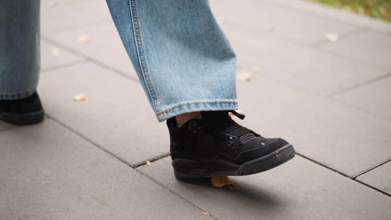 Primo piano dei piedi di una persona con sneakers nere e jeans blu che cammina su un marciapiede grigio con foglie sparse. Il soggetto si muove costantemente mostrando la trama del tessuto denim e i dettagli delle calzature.