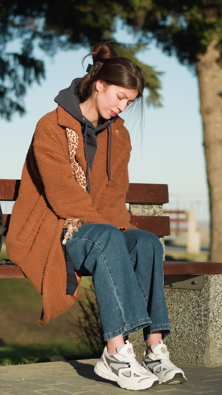 한 소녀가 도시 공원의 벤치에 혼자 앉아 있으며, 그녀의 머리는 그녀의 손에 머리를 고 깊이 우울해 보입니다. 그녀는 갈색 코트, 파란 청바지, 색 신발을 입고 있으며, 그녀의 에는 검은색 배이 있습니다.