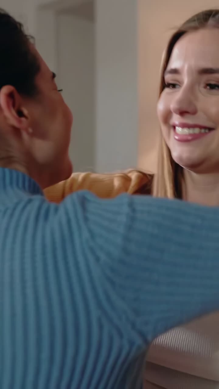 Video verticale: donna sorridente in maglia gialla che abbraccia un'amica a casa, la quale indossa una maglia blu e borchie