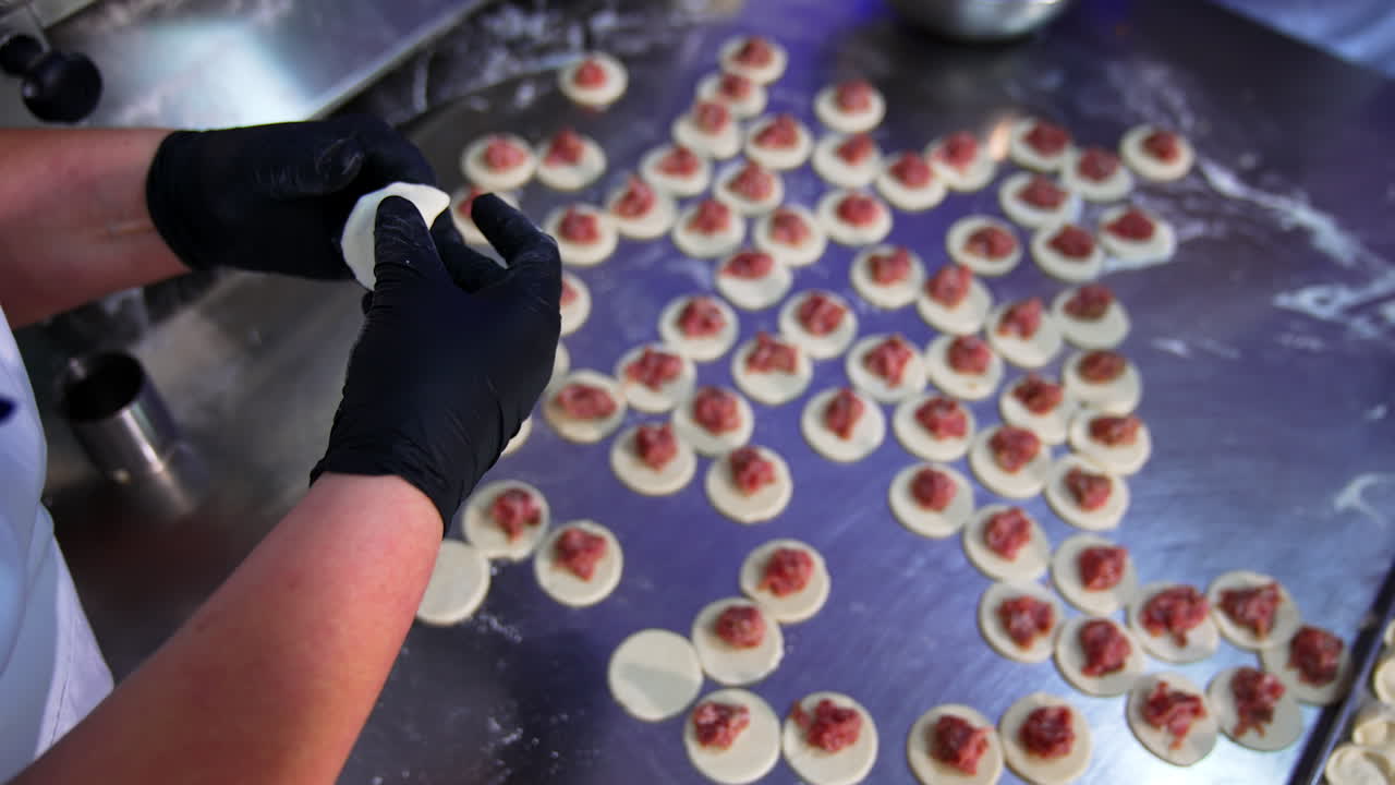 Gloved hands make ravioli. Many circles of dough with meat stuffing are placed on the metal table. View from top.