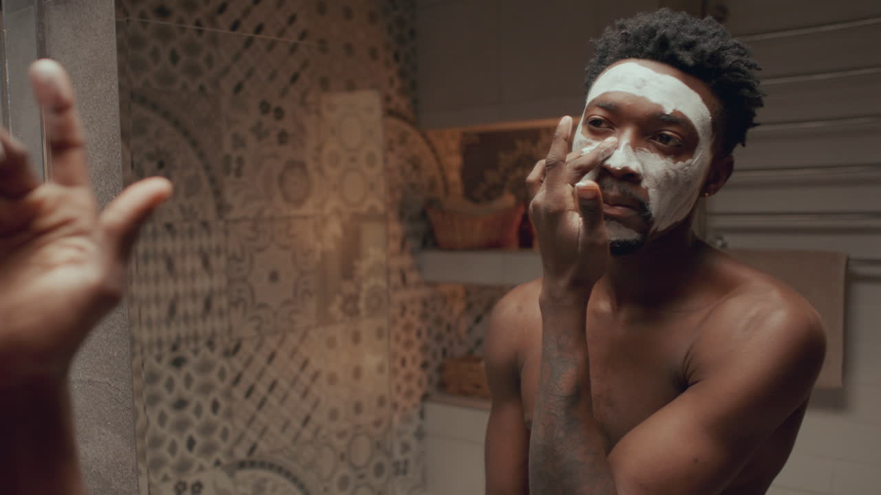 joven negro aplicando una máscara facial frente al espejo del baño