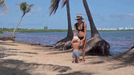 un joven niño latino siendo balanceado por su madre, ambos con sombreros en la playa en un día soleado