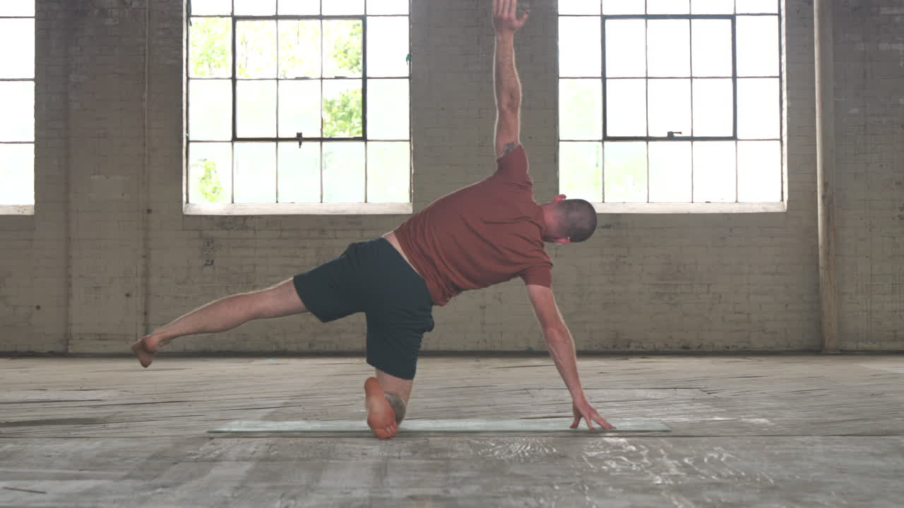 Man in an industrial warehouse practicing yoga, the pose of forward wide legged fold.