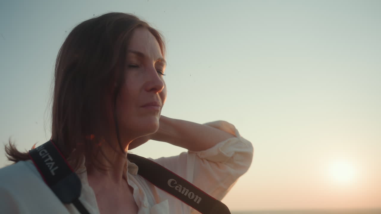 Close up side view of woman adjusting camera strap around neck with focused expression as golden sun sets behind her, soft light outlining face and hair, blurred greenery in distant background
