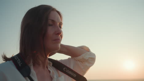 close up side view of woman adjusting camera strap around neck with focused expression as golden sun sets behind her, soft light outlining face and hair, blurred greenery in distant background