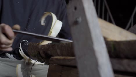 eye level shot of a carpenter peeling wood from a wooden piece to shape a chair leg with his sharp tool