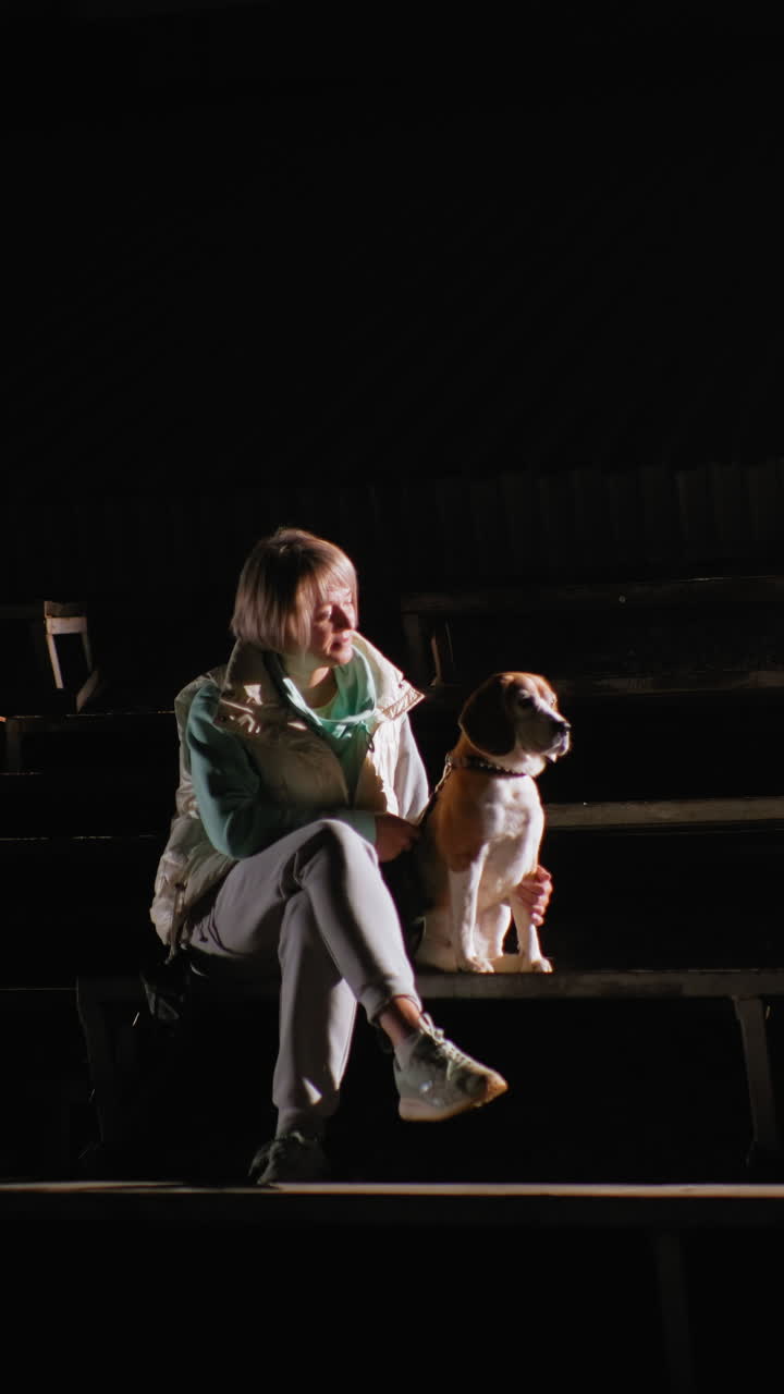 Mujer blanca sentada en las gradas bajo un único foco, con un beagle cerca, chaqueta informal y zapatillas, estadio nocturno con filas vacías, mirada contemplativa, vínculo cálido destacado por un estilo cinematográfico
