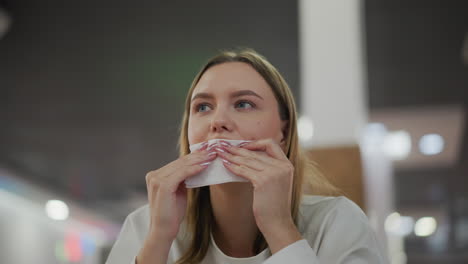 young woman sitting alone at table in mall, cleaning her mouth with tissue paper, enjoying her meal in a relaxed environment, blurry background with colorful blinking lights