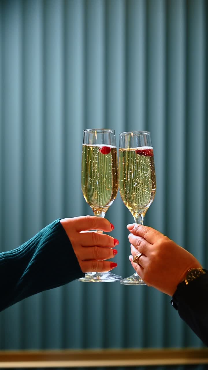Dos personas chocando copas de vino espumoso con frutos rojos. Primer plano de dos manos sosteniendo copas de champán con una sola baya en su interior sobre un fondo texturizado de color verde azulado oscuro.